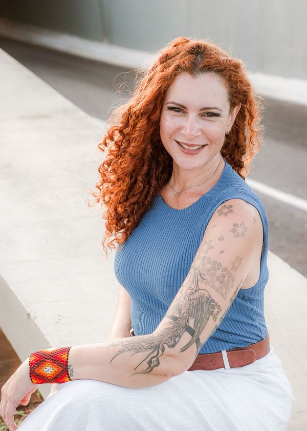 Jaya Roberta sentada, sorrindo, de blusa azul e pulseira bordada, em Brasília